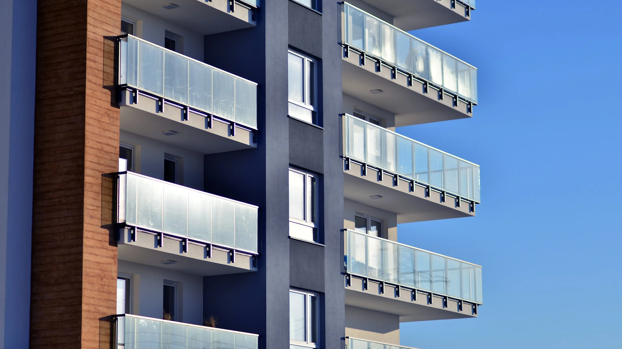 A modern multi-story apartment building with balconies featuring glass railings, a brown brick section on the side, and a clear blue sky in the background.