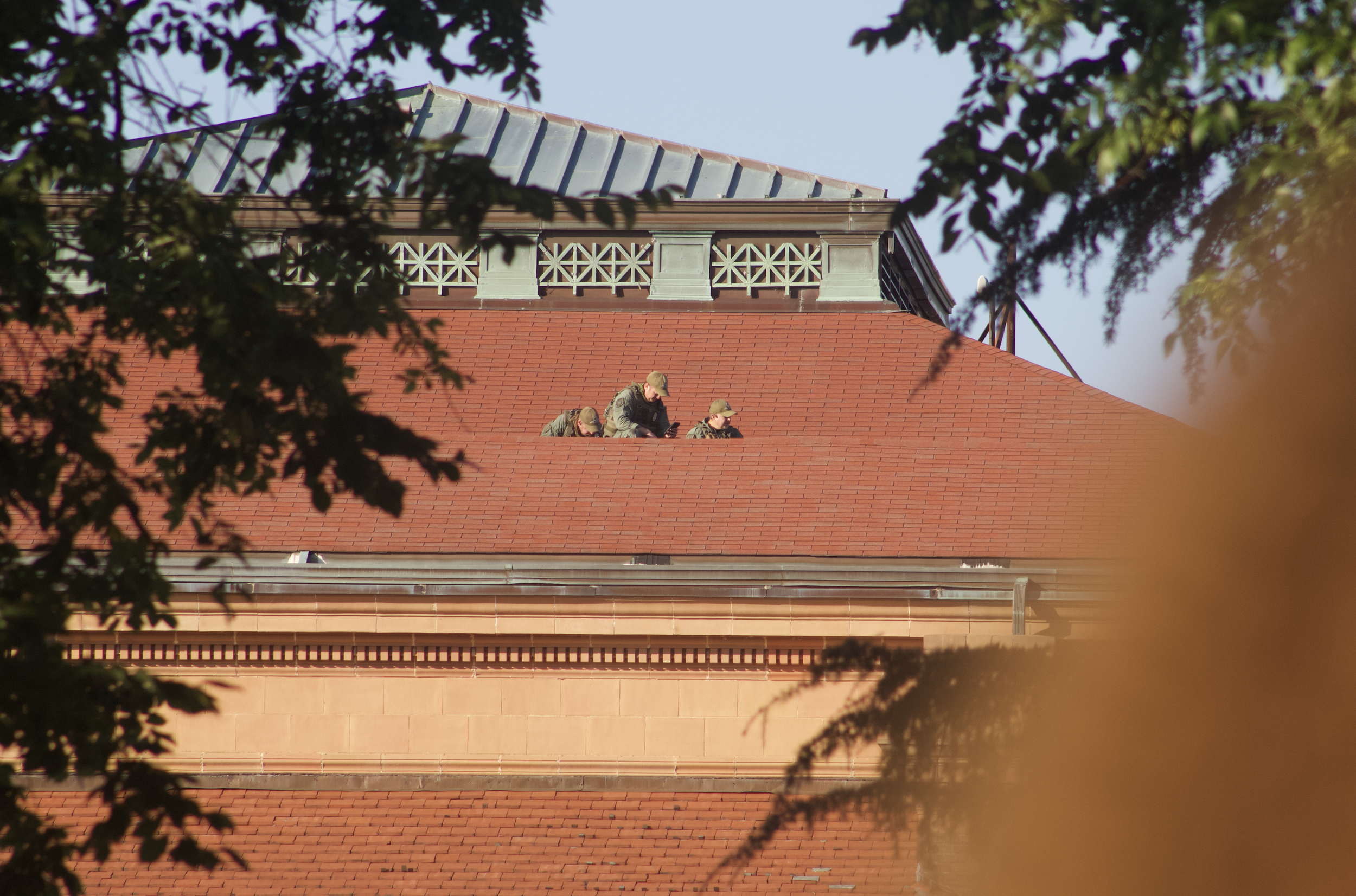Three soldiers wearing military uniforms and hats sitting on the roof of a building with red tiles, surrounded by green trees.