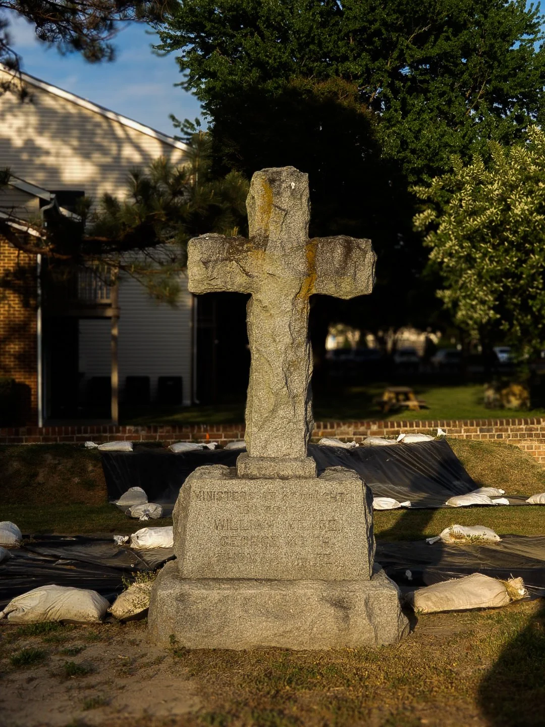 A stone cross monument in a park with trees and a building in the background, surrounded by sandbags and black material.