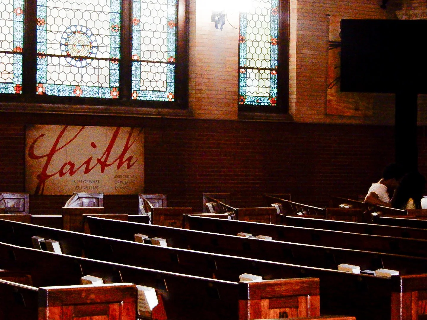 Interior of a church with stained glass windows, wooden pews, and a sign that reads 'Faith' on the wall. Two people are sitting in the pews on the right side.