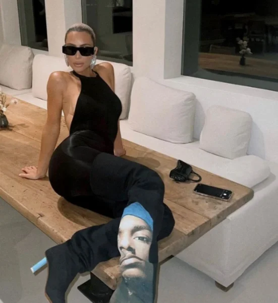 Woman in black dress and sunglasses sitting on a wooden table with her leg extended over a man wearing a hoodie with a FaceTime screenshot printed on it.