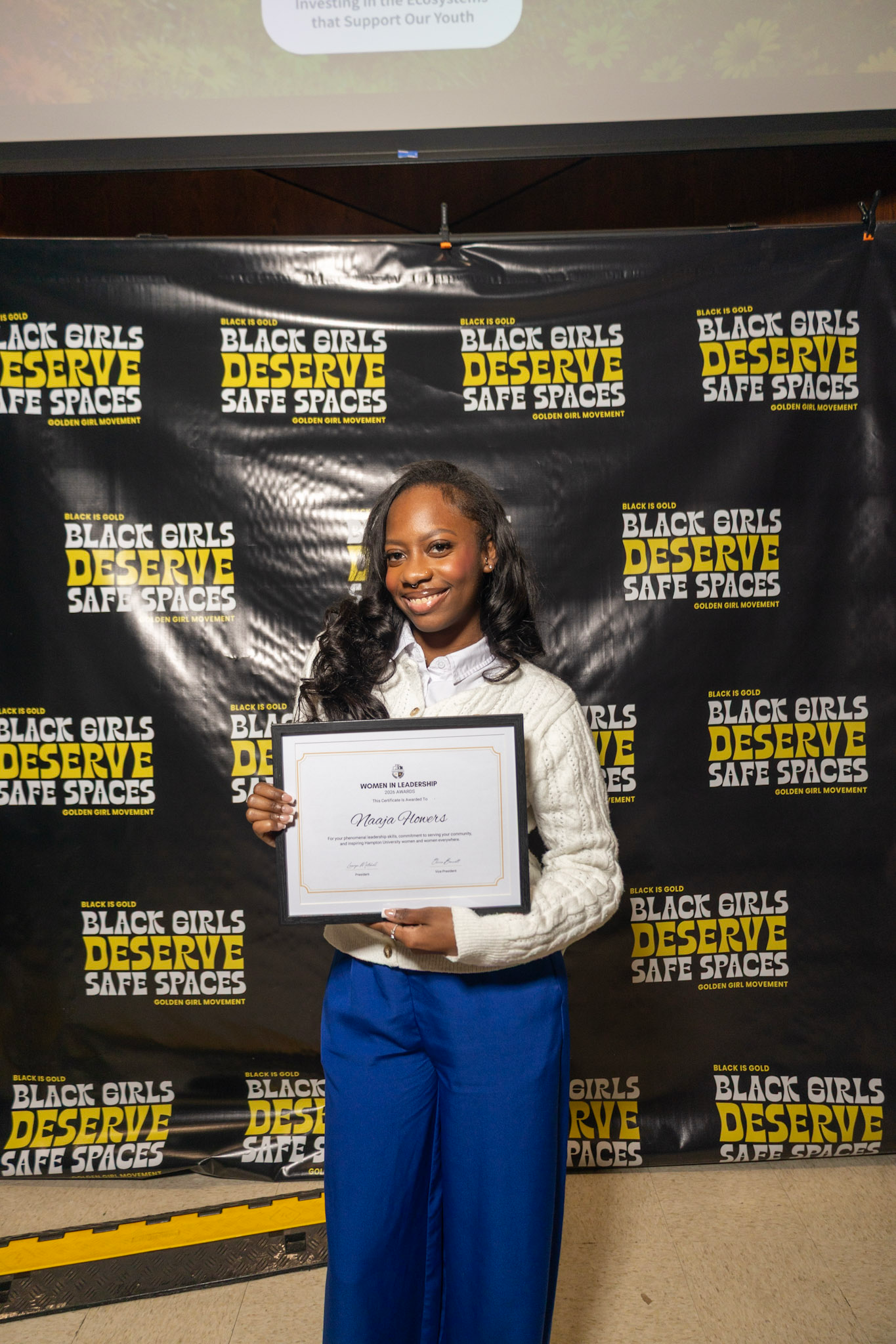A young woman dressed in a white sweater and blue pants, holding a certificate, standing in front of a backdrop with the phrase 'Black Girls Deserve Safe Spaces' repeated multiple times in yellow and white text on a black background.