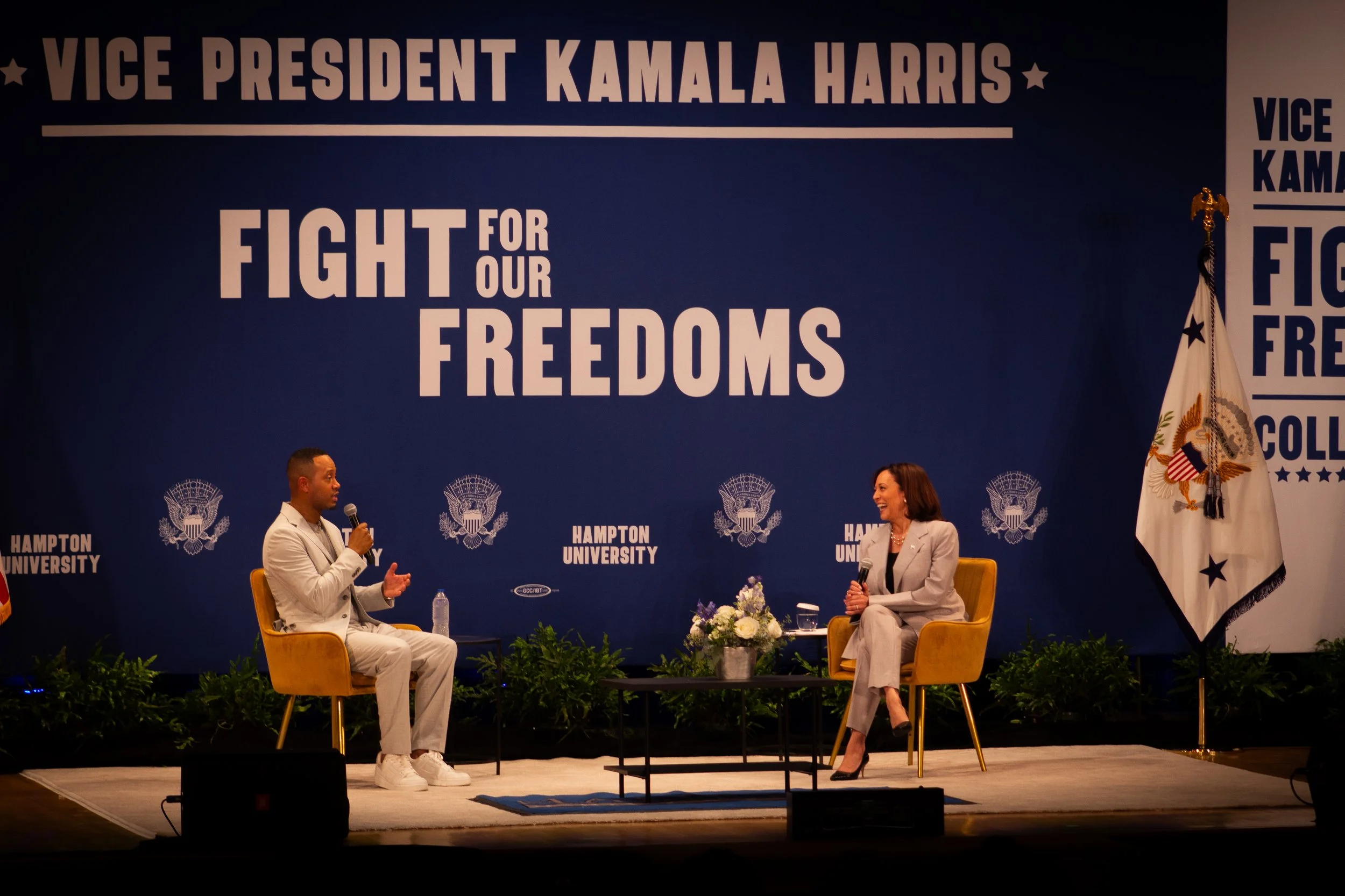 Two people speaking on a stage with a blue backdrop reading 'Vice President Kamala Harris' and 'Fight for Our Freedoms.' A woman in a light-colored suit with short hair and a man in a light-colored suit with a microphone.