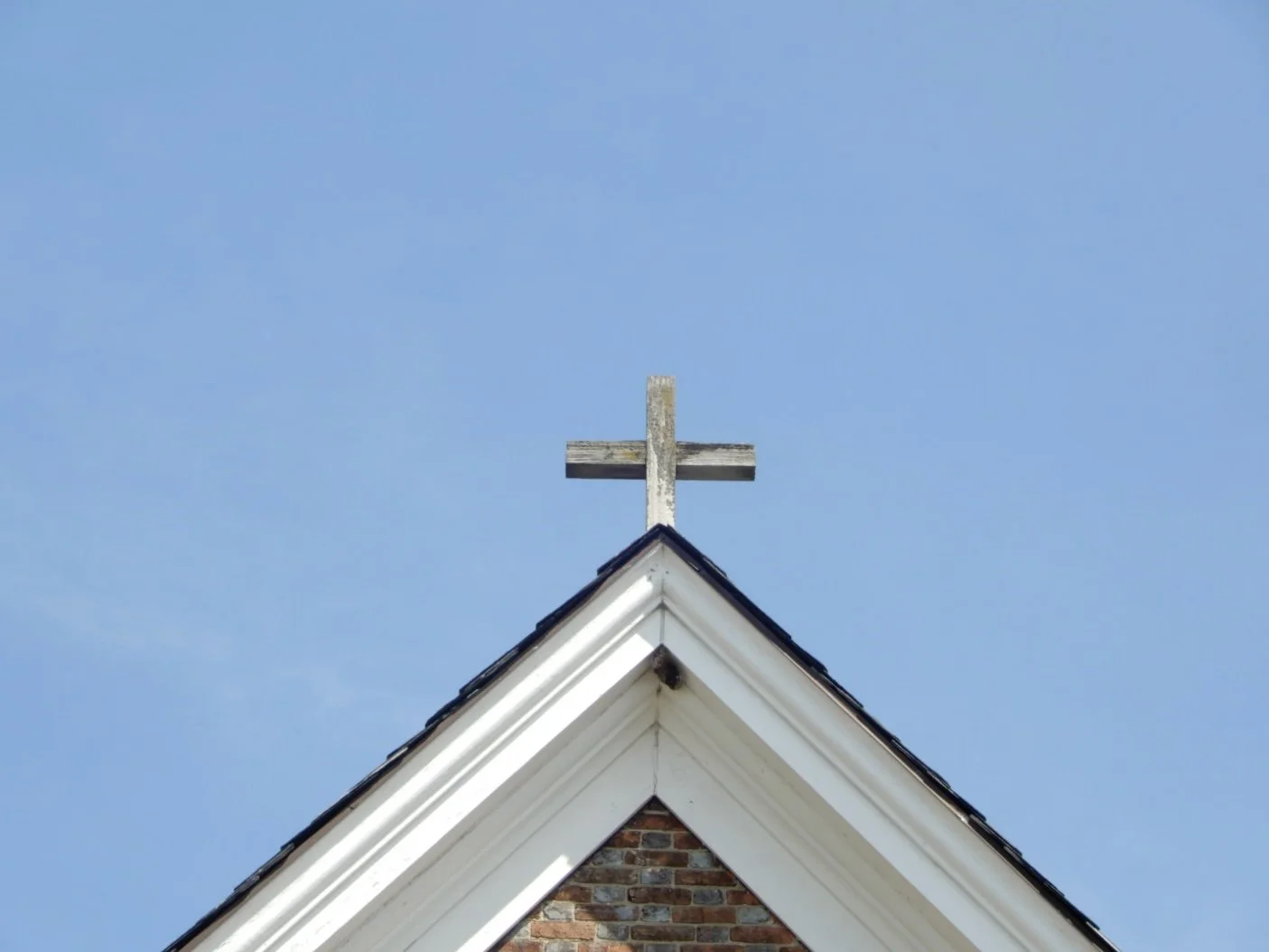 The top of a church roof with a wooden cross on the peak against a blue sky.