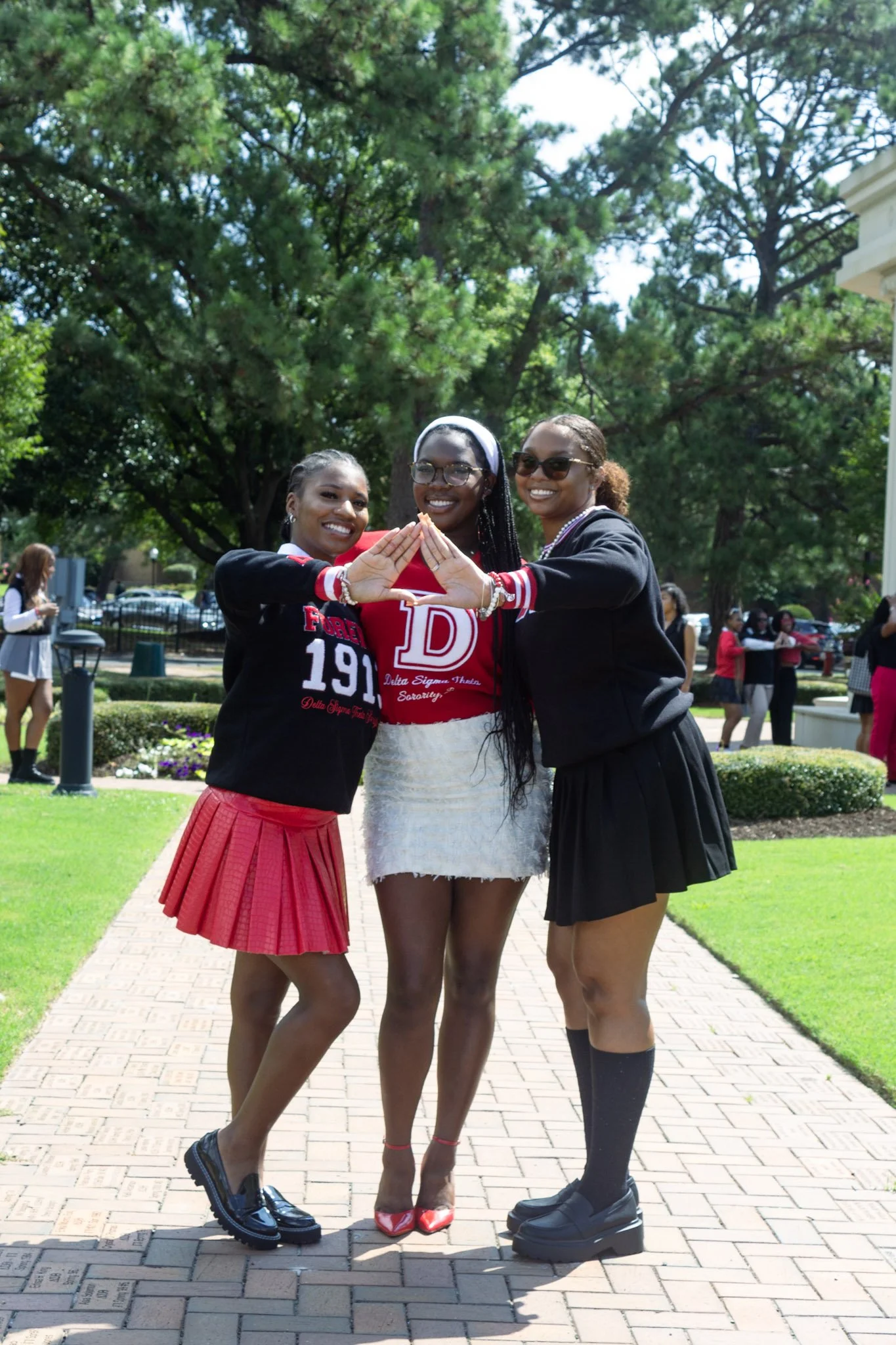 Three women standing outdoors on a brick pathway, smiling, with large trees and other people in the background. They are holding hands together in front of them.