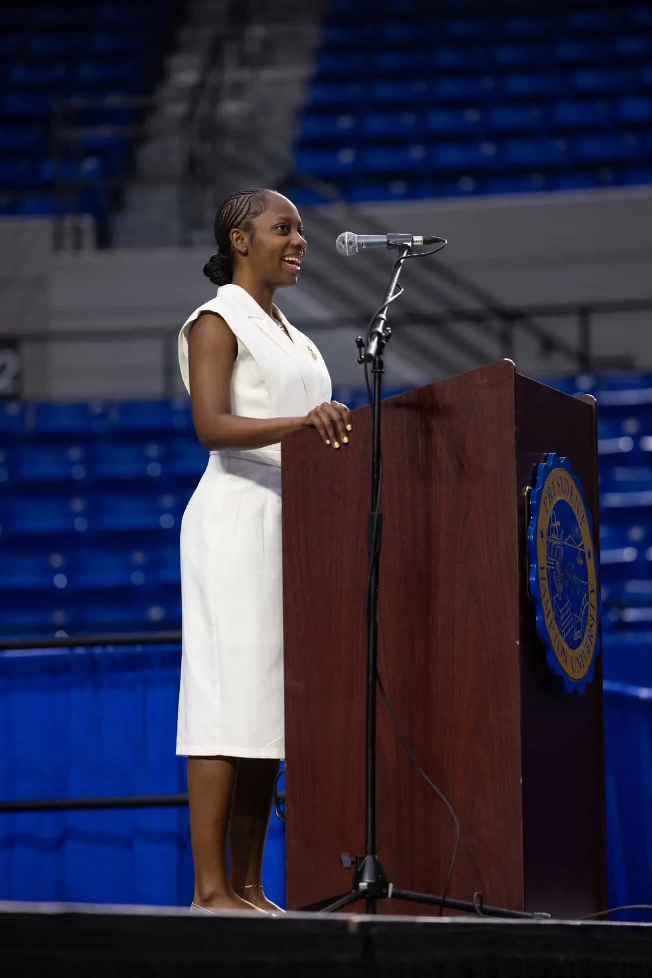 A woman in a white dress speaking at a podium in an empty stadium or auditorium filled with blue seats.