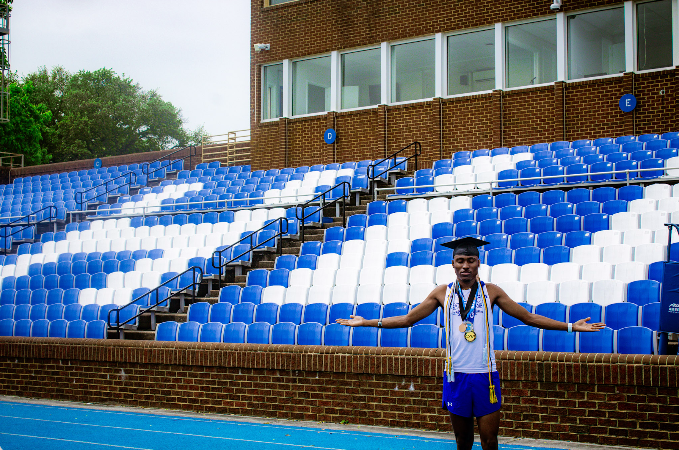 A young male athlete stands on a blue running track in a sports stadium with his arms outstretched, wearing medals around his neck and a white athletic shirt and blue shorts, with a brick building and empty blue and white seating in the background.