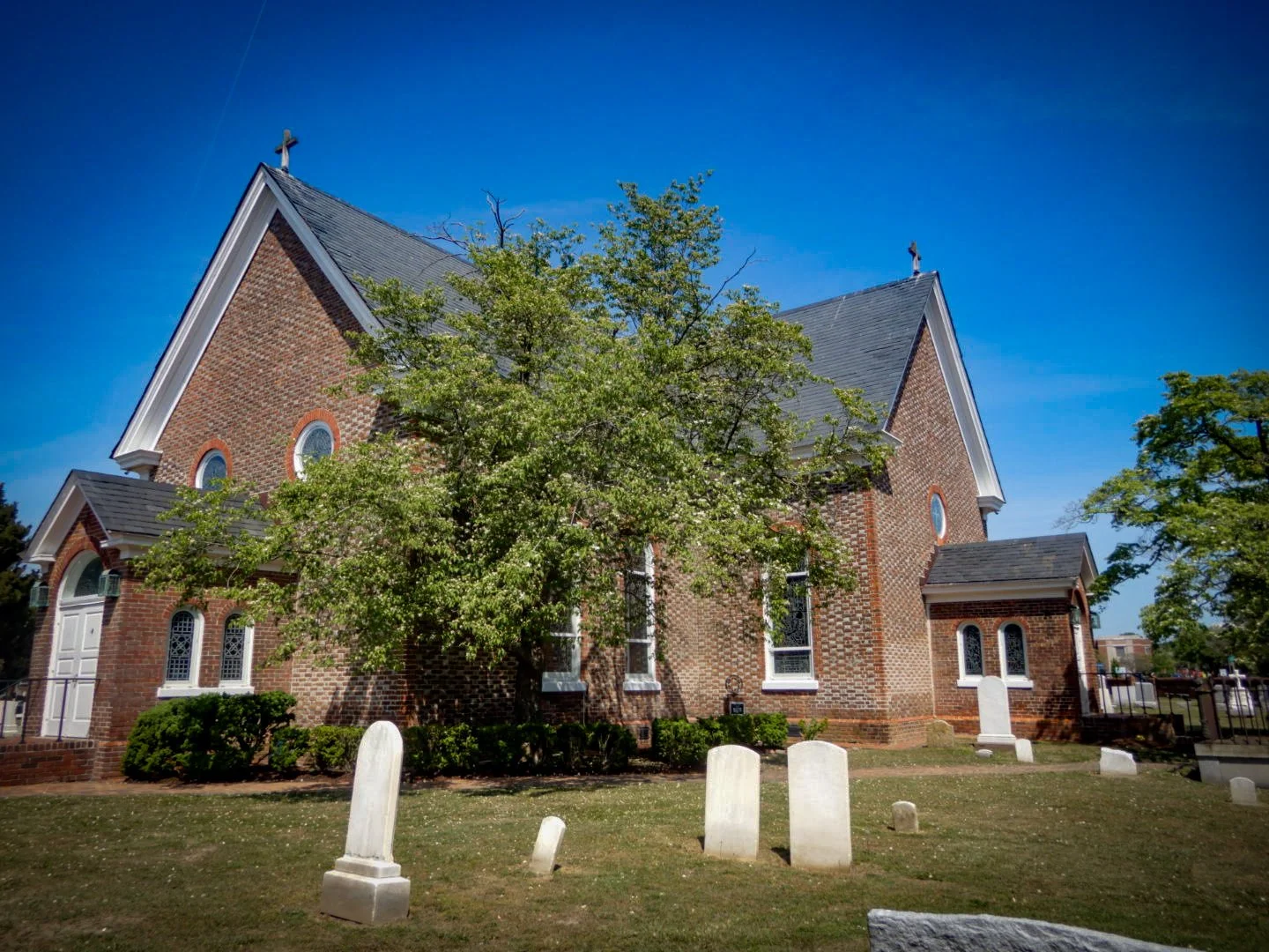 A large brick church with a steep gray roof and a cross on top, surrounded by green trees and graves in a cemetery, under a clear blue sky.