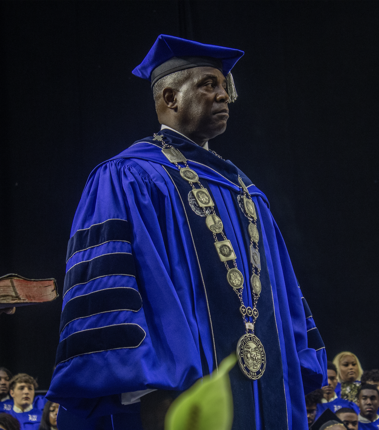 A university graduation ceremony with a man in a blue academic gown and cap, wearing a ceremonial chain, standing on stage during the ceremony.