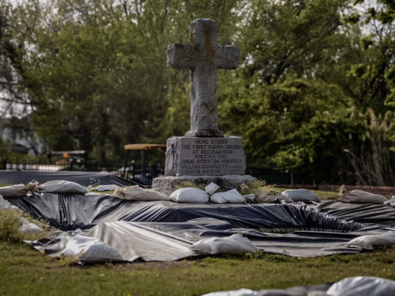 Stone cross monument with a plaque, surrounded by sandbags, outdoor setting with trees in the background.