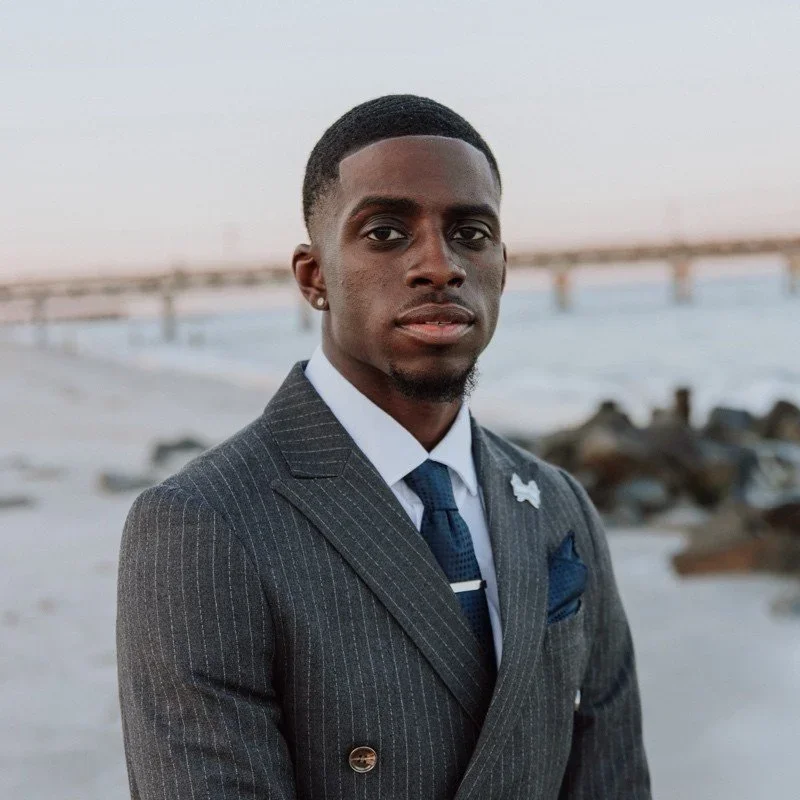 A young man in a pinstripe suit and tie standing on a beach with a pier and rocks in the background.