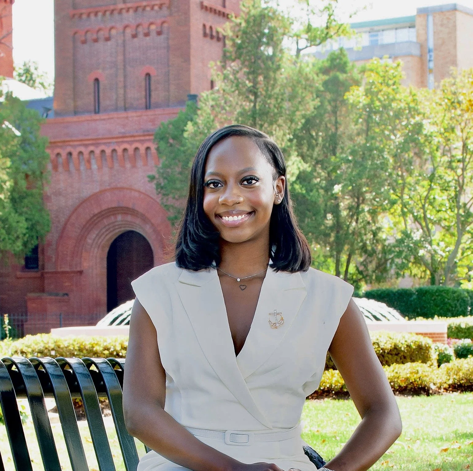 A smiling young woman with shoulder-length black hair sitting on a park bench, wearing a white sleeveless blazer with a small anchor pin and a heart-shaped necklace, in front of a red brick building surrounded by trees and greenery.