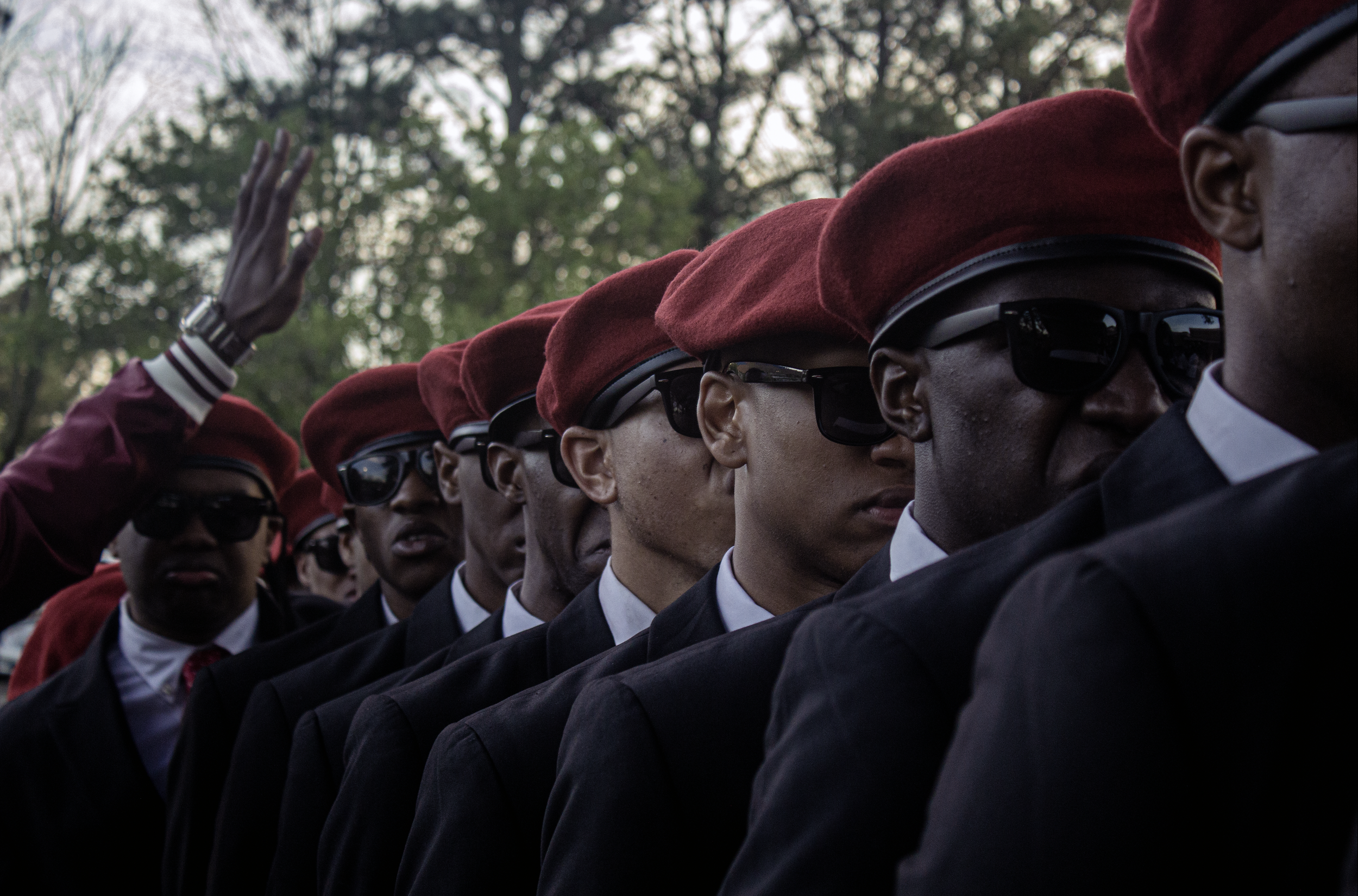 A group of individuals, wearing dark suits, white shirts, and red berets, standing in a line with their right hands raised in a salute.