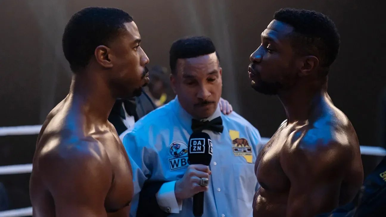 Two shirtless male boxers face each other in the boxing ring during a match, with a referee holding a microphone between them. The referee is in the background, wearing a white shirt and black bow tie, and appears to be speaking to the boxers.