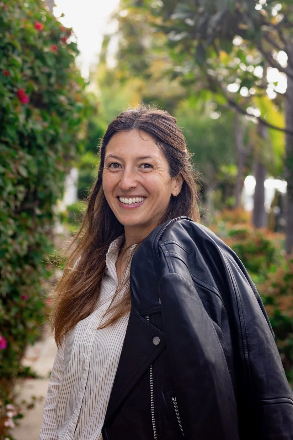 A smiling woman with long brown hair, wearing a black leather jacket over a white striped shirt, standing outdoors with greenery and plants in the background.