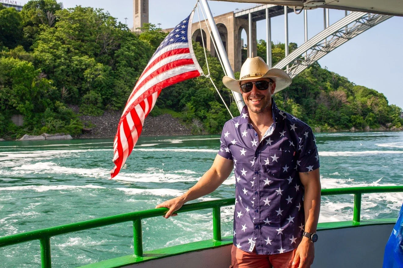 A man wearing a cowboy hat, sunglasses, and a star-patterned shirt stands on a boat holding an American flag. The boat is on a river with green hills and a large bridge in the background.