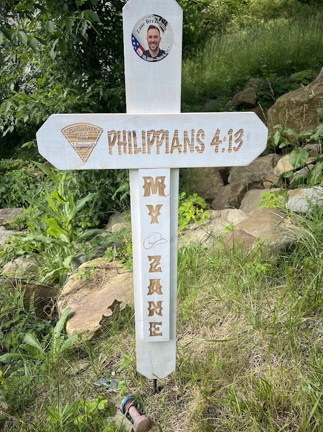 White wooden cross with engraved and painted words, surrounded by green plants and rocks. The top of the cross has a round sticker with a man's photo and text, and a triangle-shaped sticker with the text 'Morgantown Police Dept. Z. Breakiron, KY.' The cross has the words 'PHILIPPIANS 4:13' and the phrase 'M.Y. ZANE' with a small hat graphic.