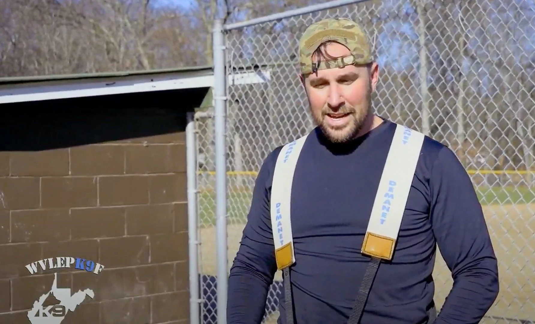 A man wearing a camouflage cap and a black long sleeve shirt, with a harness over his shoulders, standing outdoors near a chain-link fence. Zane Breakiron Uniontown Pa