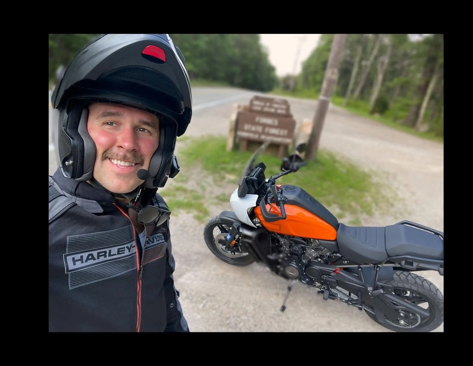A smiling man wearing a Harley Davidson jacket and helmet takes a selfie with an orange and black motorcycle on the side of a rural road, with trees and a sign in the background.