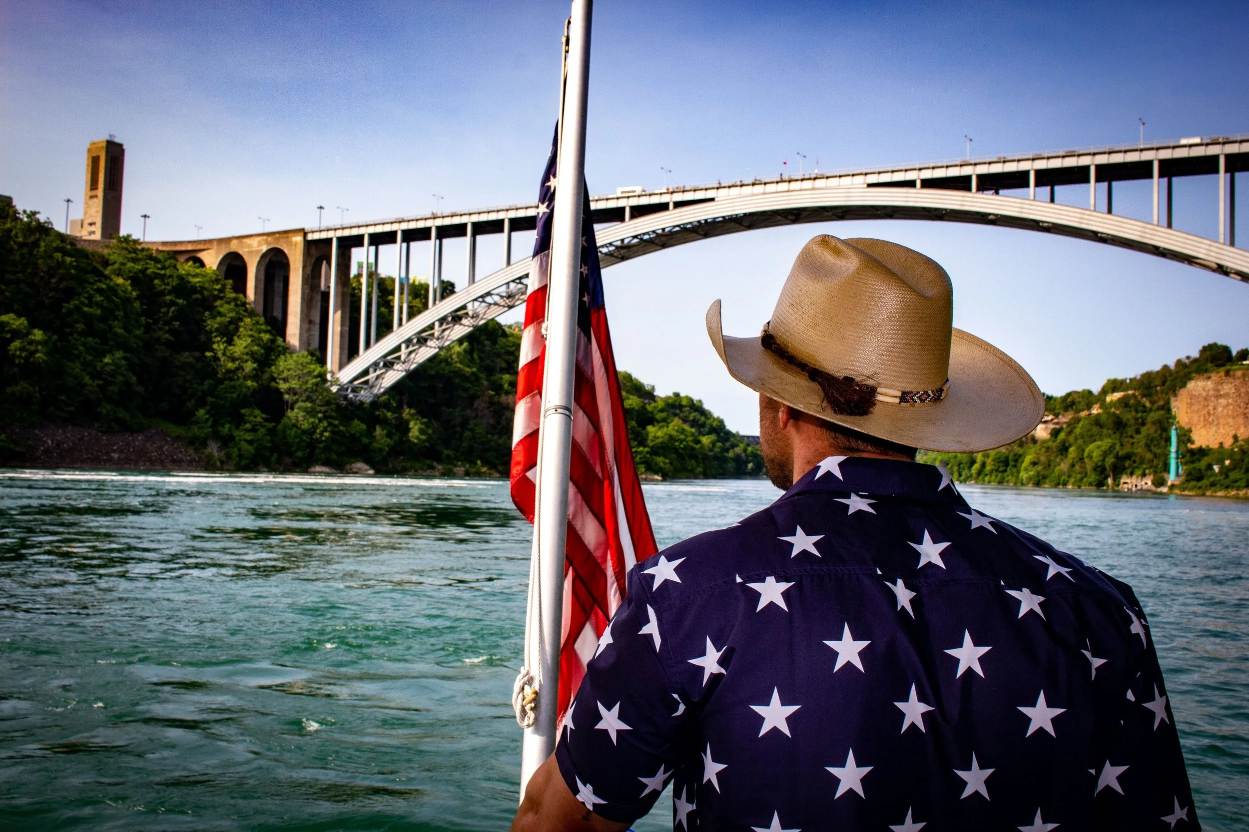 A man wearing a cowboy hat and an American flag-themed shirt, holding an American flag, is on a boat on a river near a large bridge with greenery and a tower in the background. Zane Breakiron Uniontown Pa