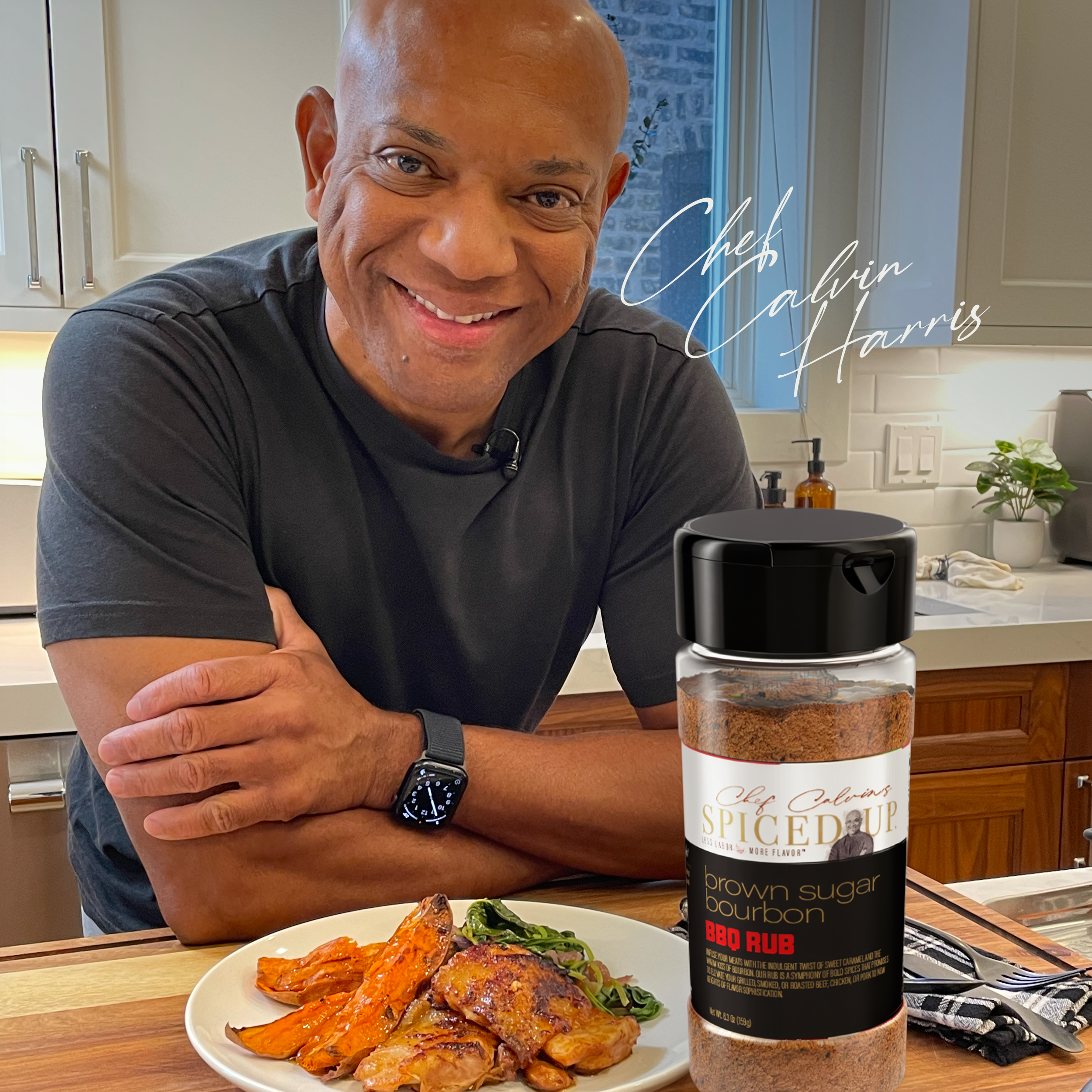 A man smiling at the camera sitting at a kitchen table with a plate of cooked food, including roasted vegetables and meat, in front of him. There is a jar of brown sugar bourbon BBQ rub on the table and a window in the background.