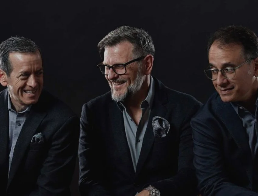 Three men in business suits are sitting together, smiling and looking at each other against a black background.