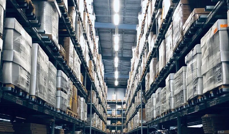 A warehouse aisle with tall shelves stocked with boxes on both sides, extending into the distance under bright overhead lighting.