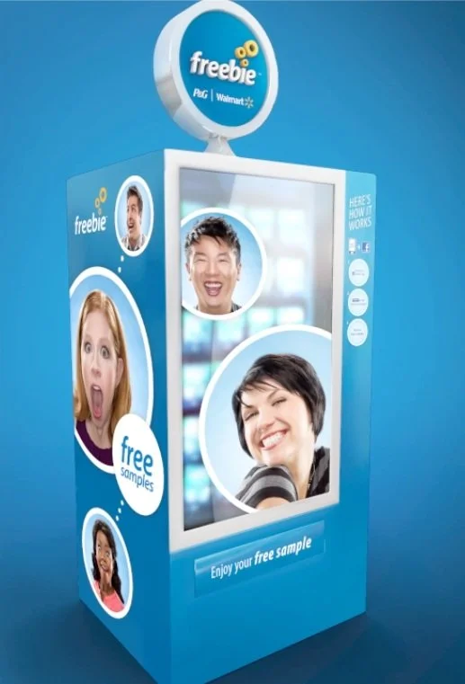Kiosk vending machine offering free samples and promotional materials with photos of diverse happy people, branding for Freebie, P&G, and Walmart, set against a blue background.