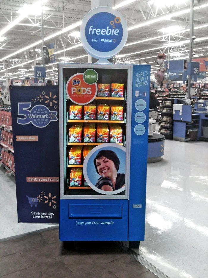 A vending machine inside Walmart offering free samples of Tide Pods, with branding and promotional signage indicating it's a new product, and a smiling woman on the display.