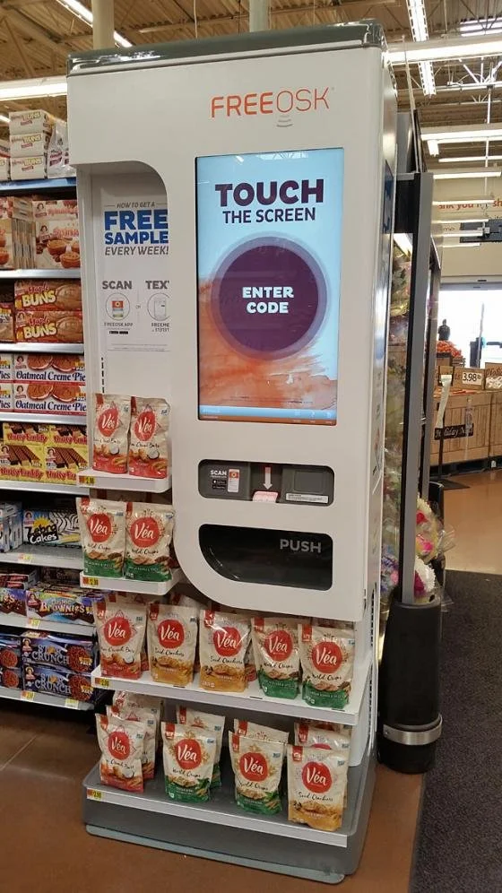 Self-serve sample kiosk at a grocery store with a screen that says 'Touch the Screen' and 'Enter Code,' a scanner, and a push button, with bags of Vea pancake mix on display below.