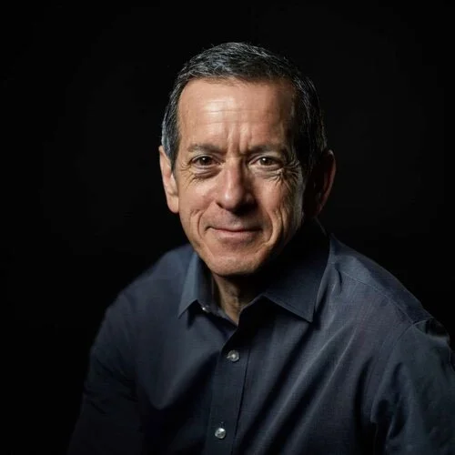 Portrait of a middle-aged man with short dark hair, wearing a dark button-up shirt, against a black background.