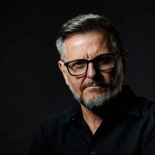 Portrait of a middle-aged man with glasses, a beard, and gray hair, wearing a black shirt against a dark background.