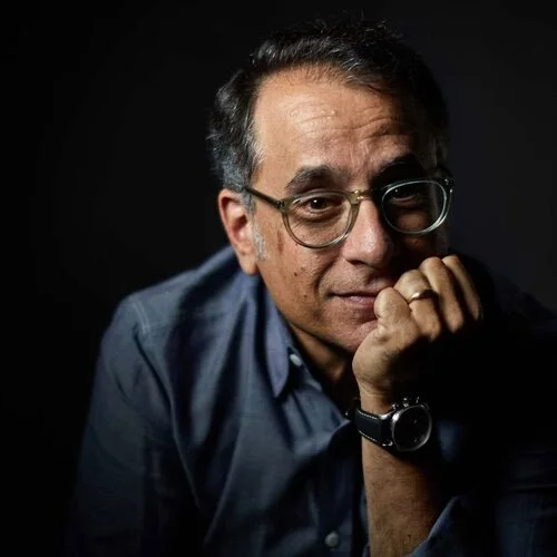 A man with glasses and dark hair, wearing a dark shirt, resting his chin on his hand against a black background.