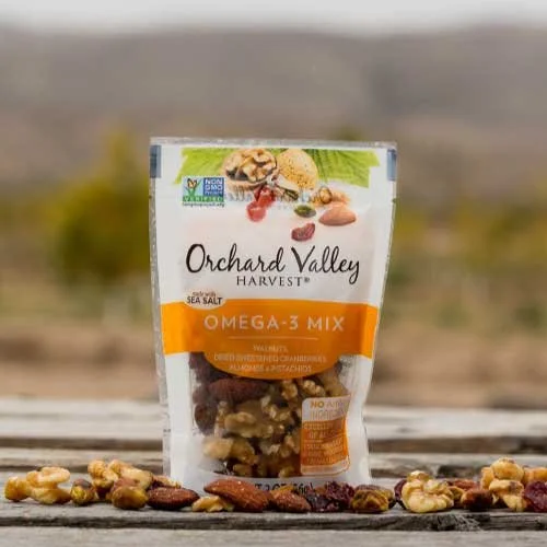 A package of Orchard Valley Harvest Omega-3 Mix trail mix on a wooden surface with loose trail mix pieces in front, outdoors with a blurred landscape background.