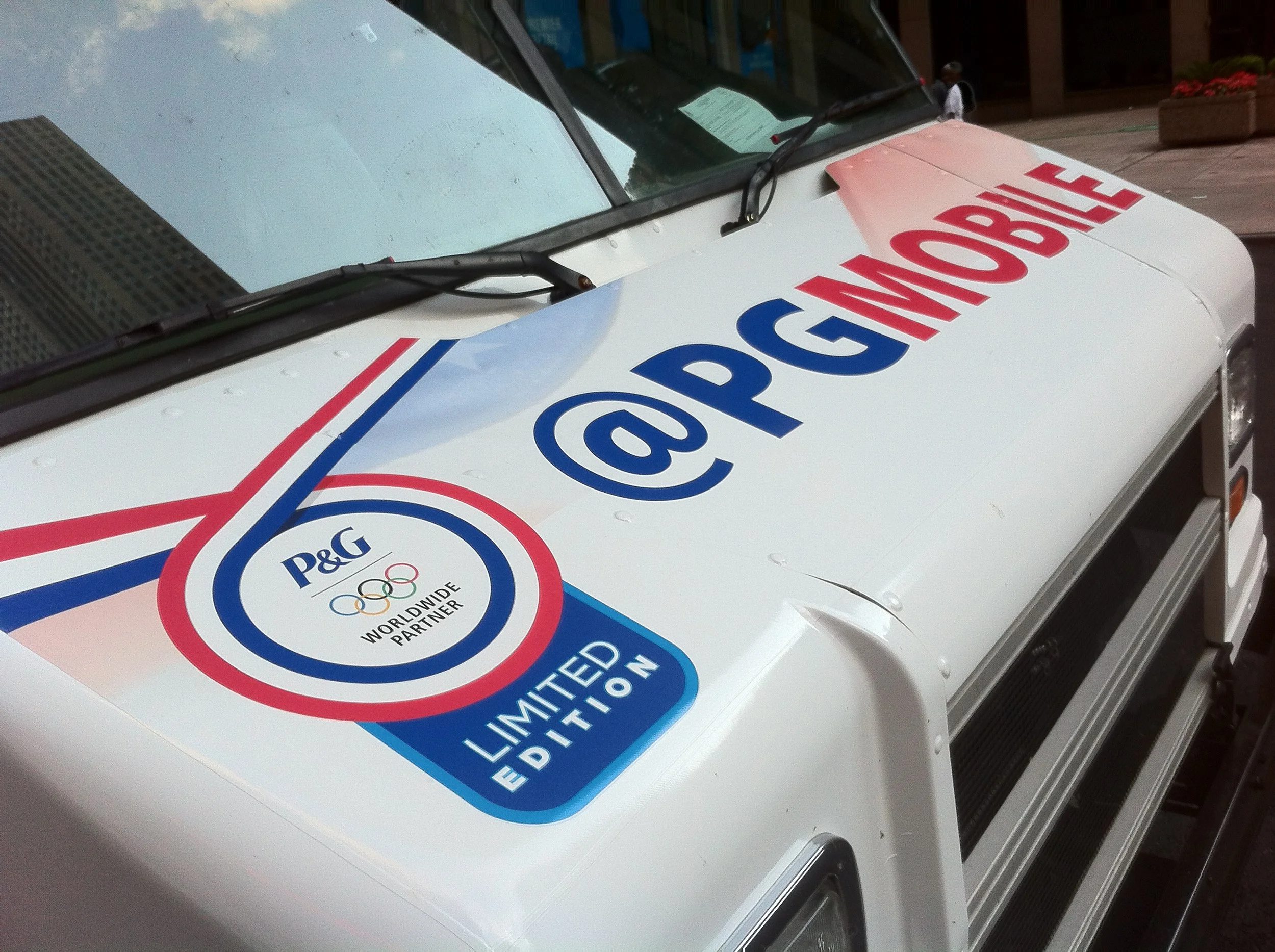Close-up of a white vehicle with a large decal reading '@PG' with the P&G logo, Olympic rings, and the words 'Worldwide Olympic Partner' and 'Limited Edition' in blue and red.