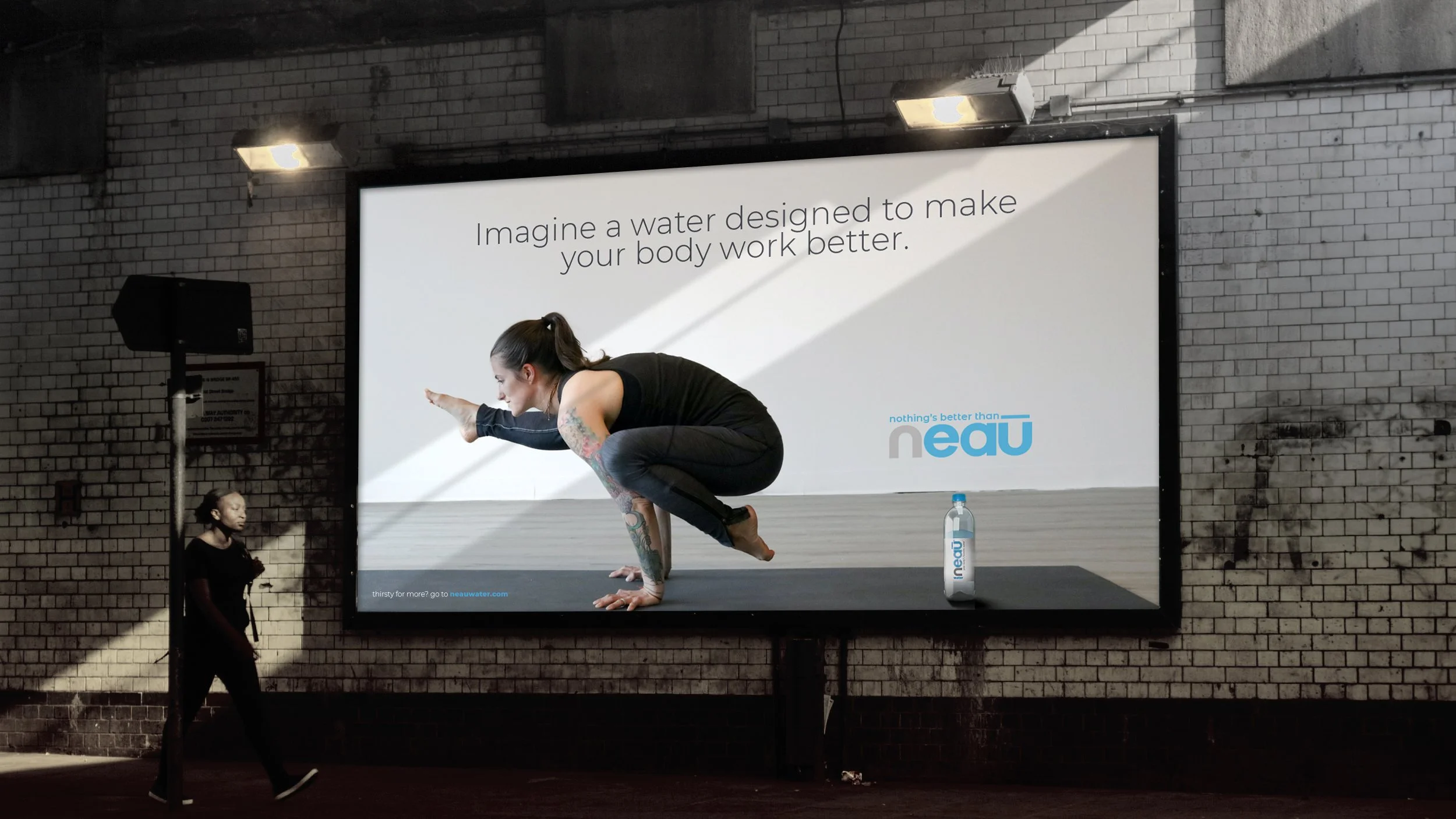 A woman with tattoos practicing yoga in front of a large advertisement for neutr water, with a bottle of neutr water on the ground nearby. There is a woman standing near the wall, looking downward.