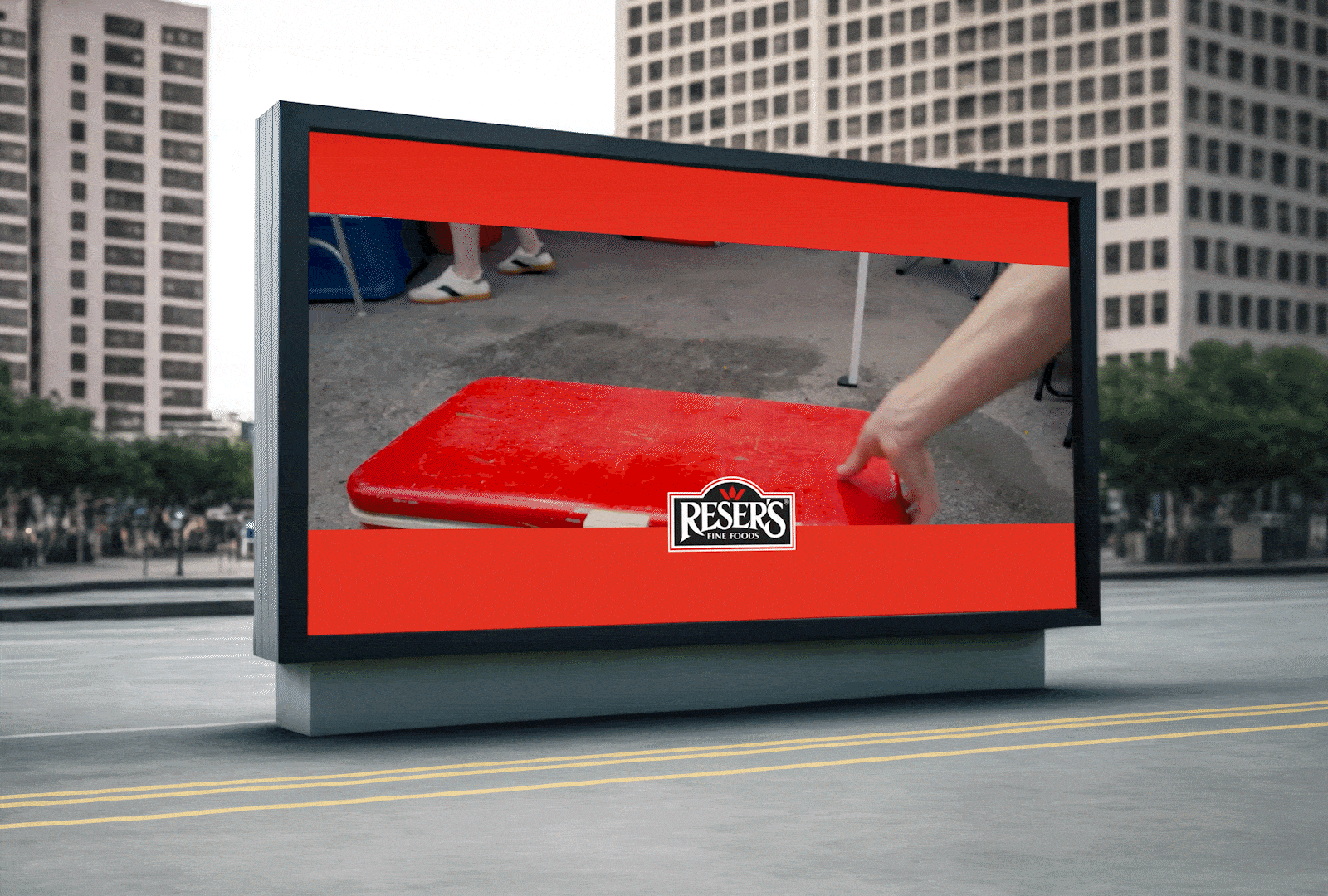 A large outdoor screen displays an image of a red cooler being placed on a surface with the logo 'Resers Fine Foods' in the foreground. The outdoor setting includes tall buildings and parked chairs in the background.