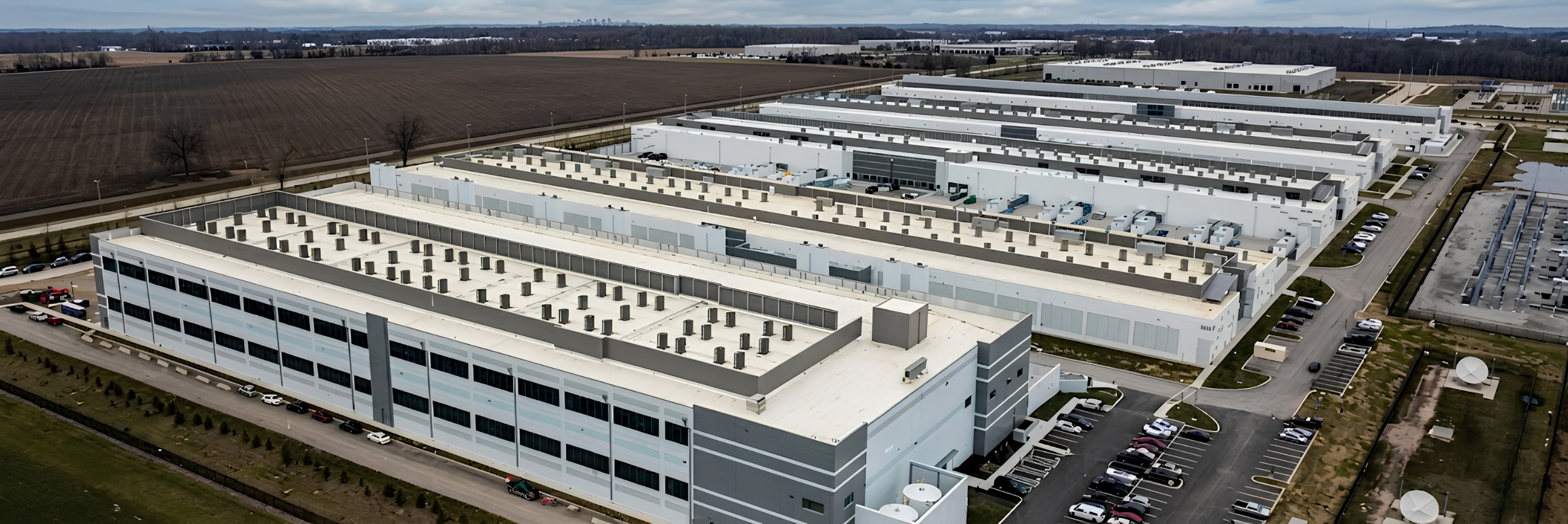 Aerial view of a large industrial or office complex with multiple white buildings, parking lots filled with cars, and surrounding open fields under a partly cloudy sky.