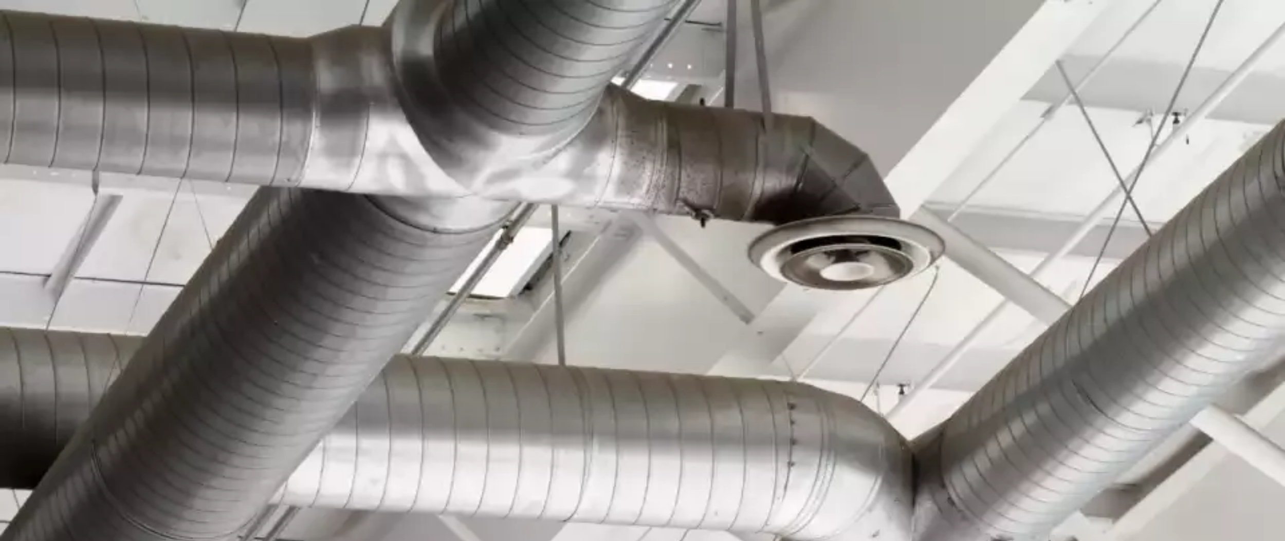 The image shows several large silver industrial air ducts and a ceiling-mounted air vent in an indoor space with a white ceiling.