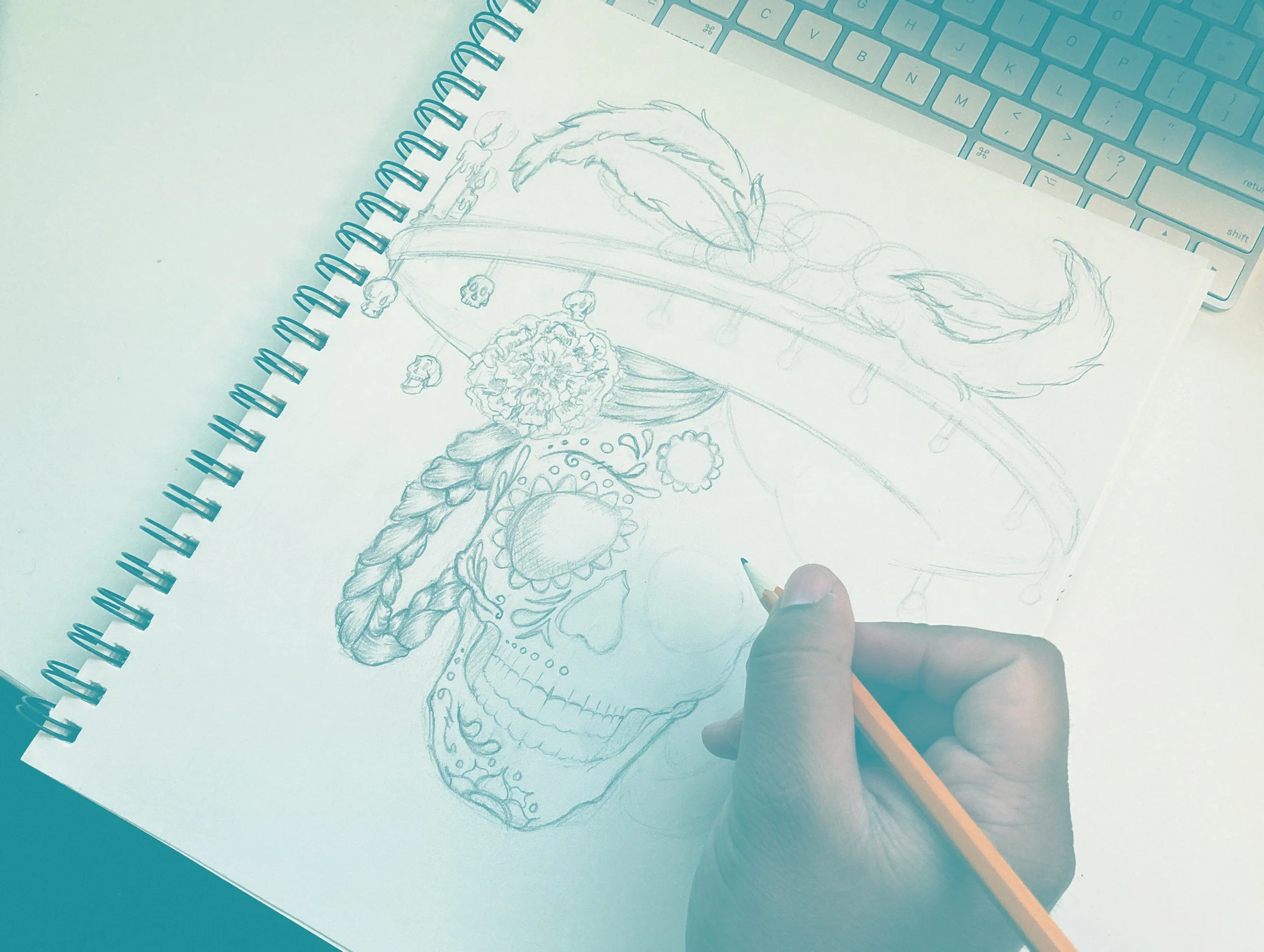 A sketch of a dragon on a spiral-bound notepad with decorative skulls hanging from a band on its neck, placed on a desk next to a blue keyboard and a person's hand holding a pencil.