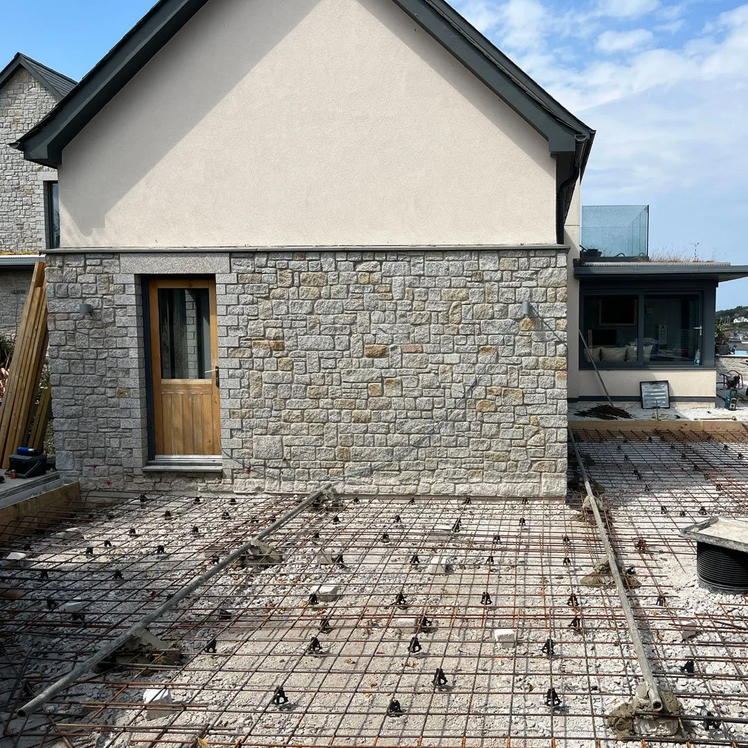 Construction site with rebar grid for concrete, partially built house with stone and stucco exterior, and a small wooden door.