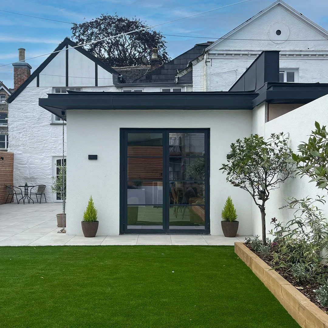 Modern house with white walls, black sliding glass doors, and black roof trim. There are two potted plants on either side of the door, a small tree in a flower bed, and a lush green lawn in the foreground. An outdoor seating area with two chairs and a table is visible on the left.