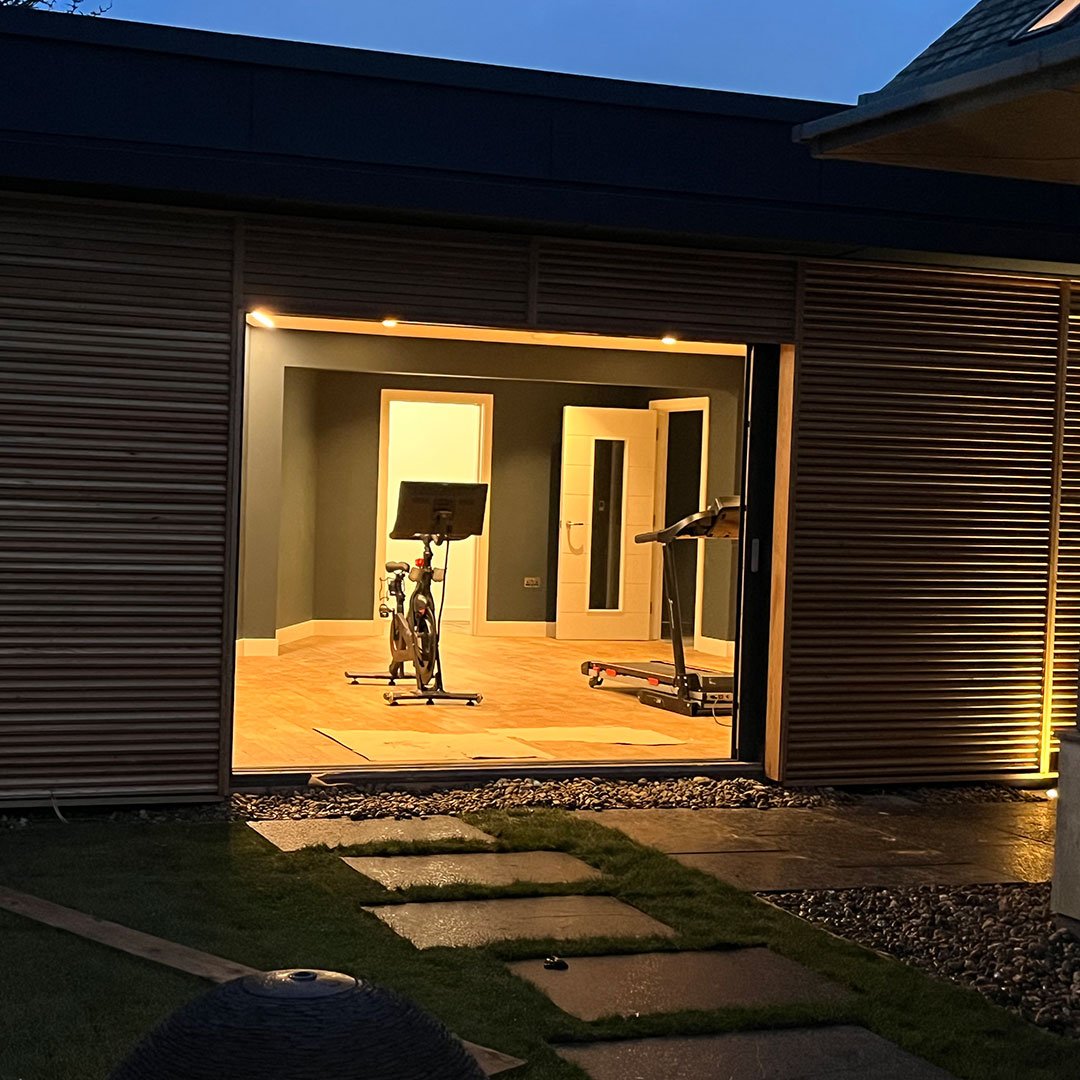 An illuminated home gym seen through a garage door, with an exercise bike and a treadmill on wooden flooring, in a house with dark walls and white trim.
