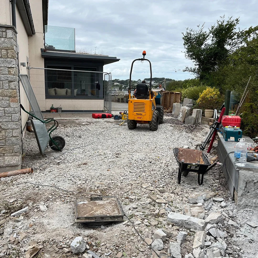 Construction site showing gravel ground, a wheelbarrow, a bulldozer, construction tools, and materials, with a house and trees in the background.
