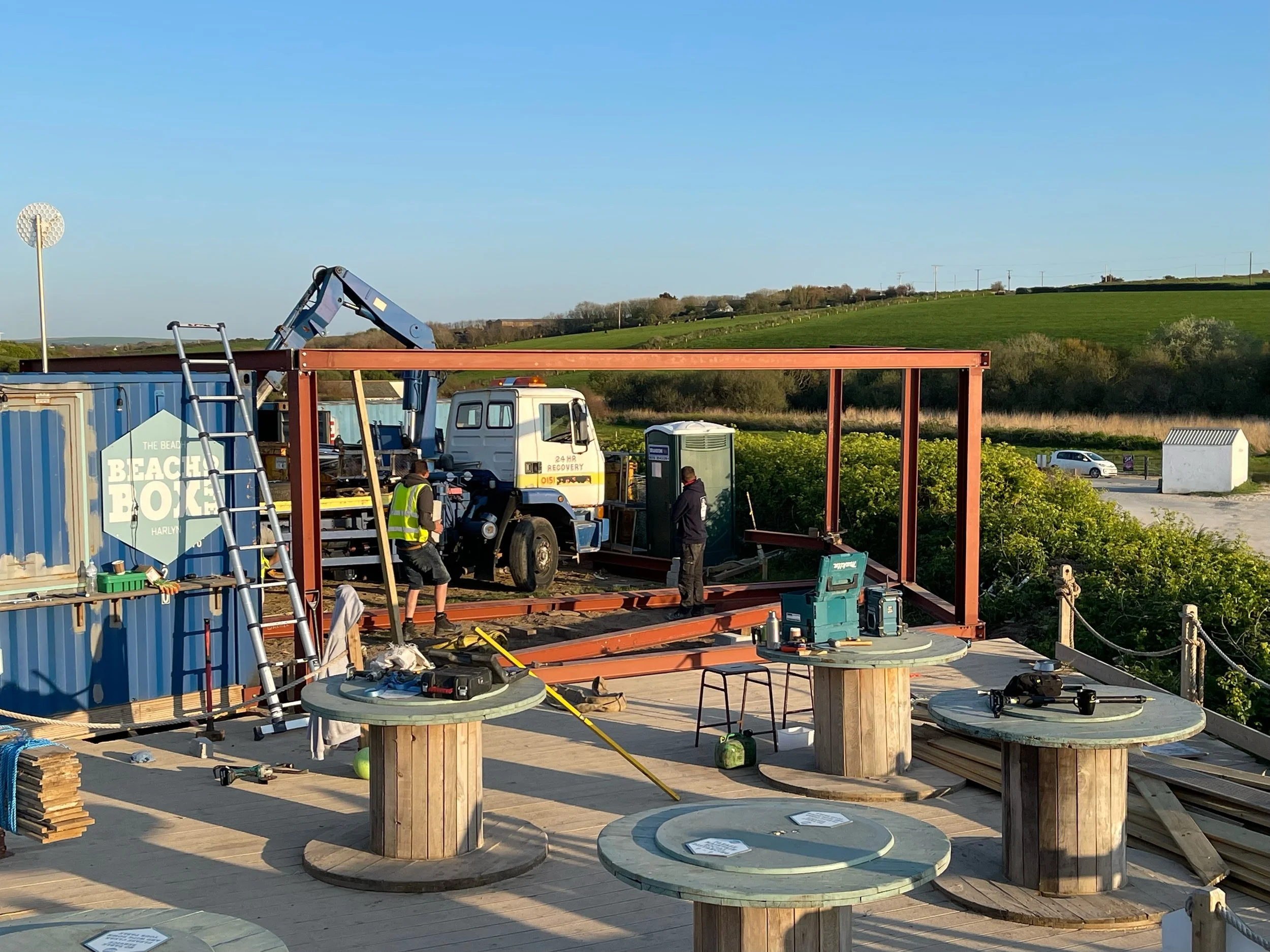 Construction site with workers assembling a metal frame structure outdoors with green hills and a blue sky in the background.