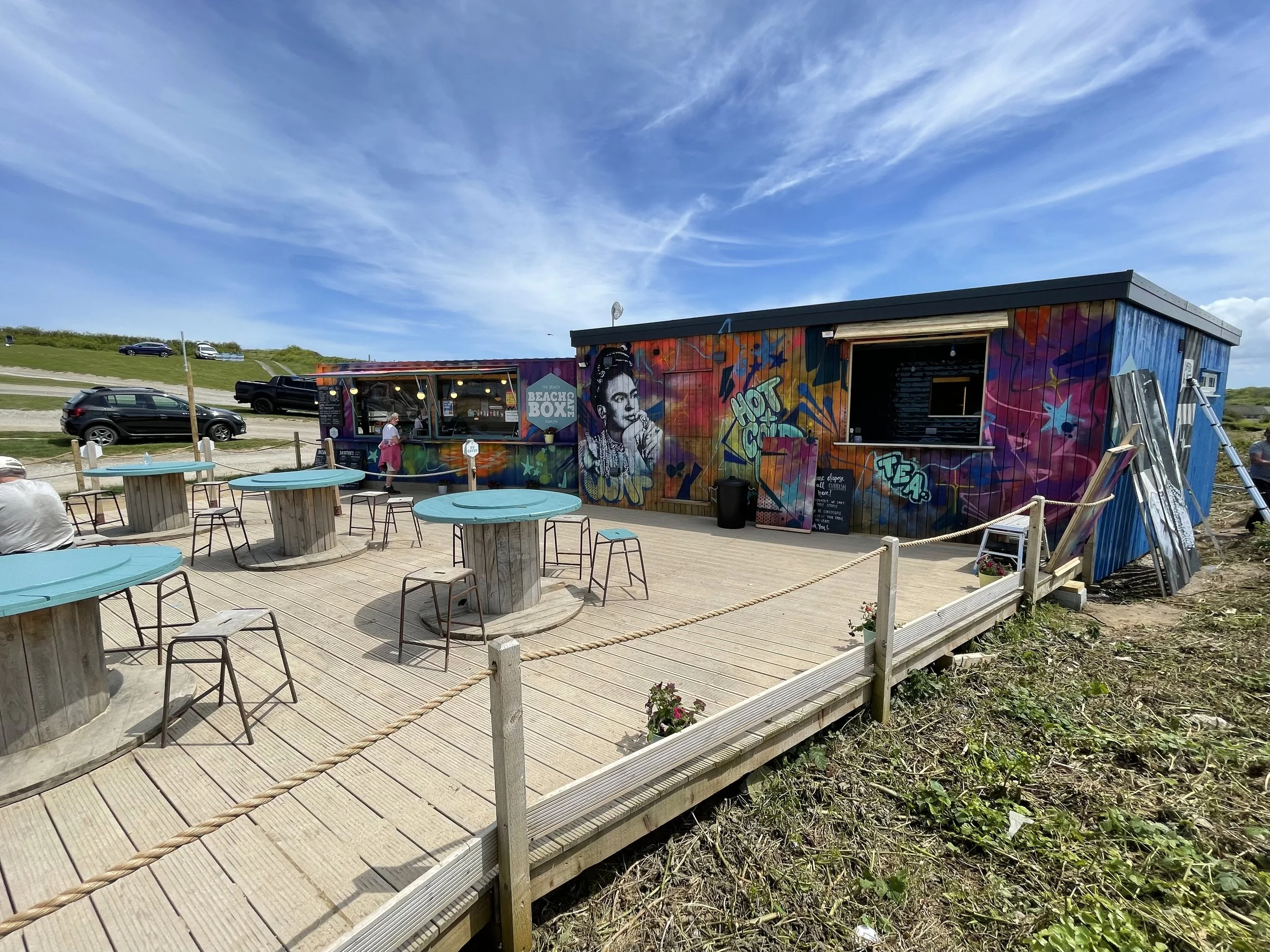Colorful graffiti-covered building with outdoor seating area and several parked cars nearby. Bright blue sky with some clouds.