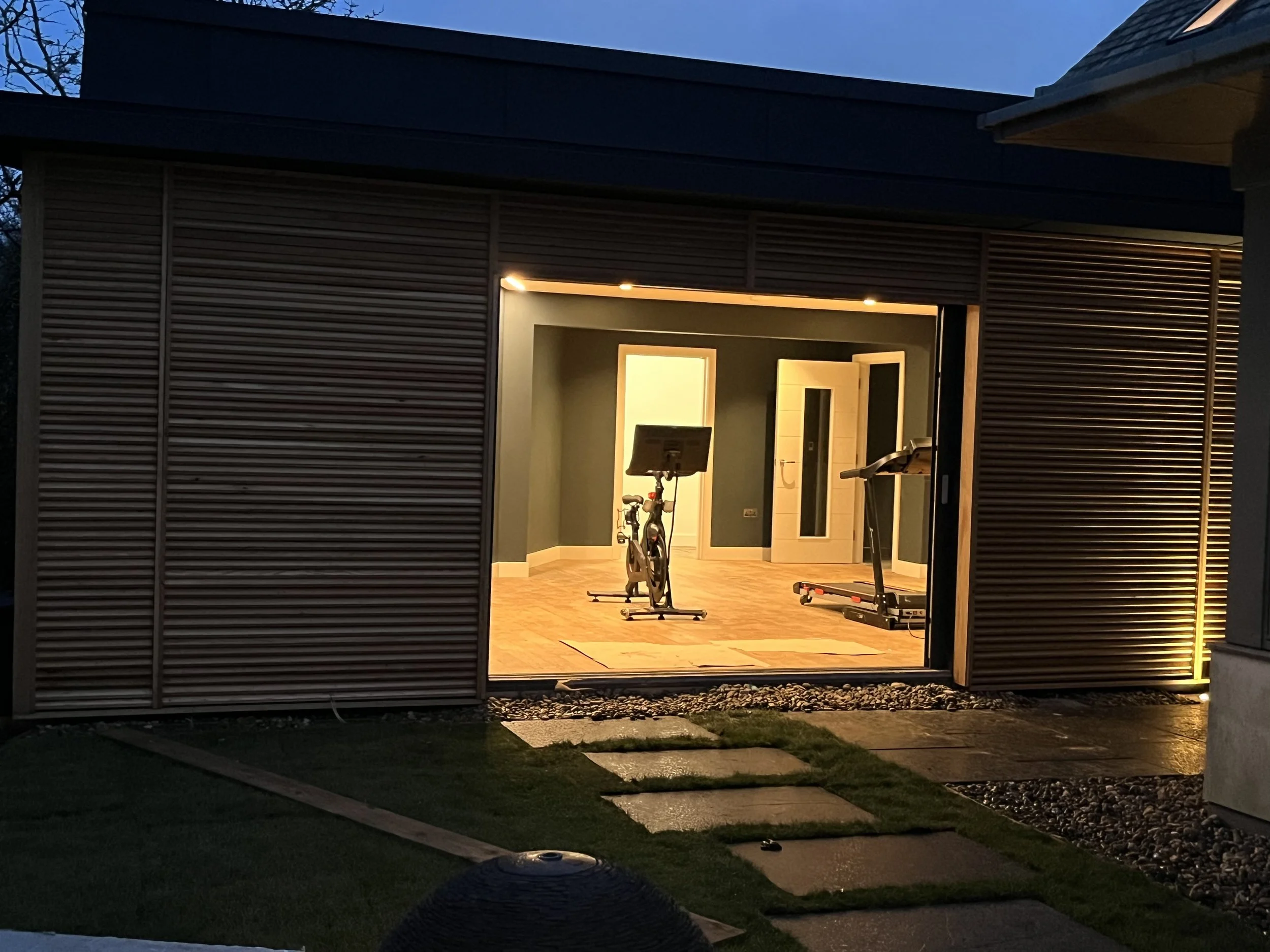 View of a garage with open doors revealing a home gym with exercise equipment, including a stationary bike and treadmill, illuminated by interior lighting during dusk.