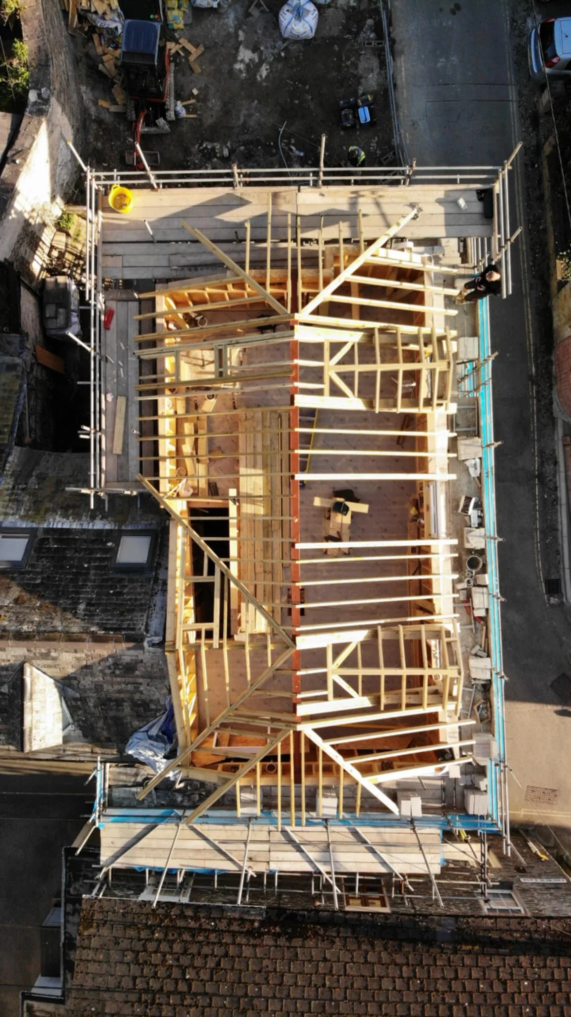 Aerial view of a house under construction with wooden framing and scaffolding, surrounded by vehicles and construction materials.