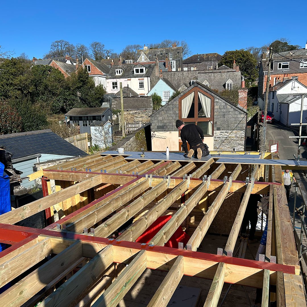 Construction workers working on a building frame with wooden beams on a rooftop in a neighborhood with houses and trees.