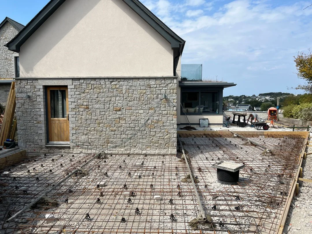 Construction site showing reinforced steel rebar on the ground for a concrete slab in progress, next to a house with stone and stucco exterior walls.